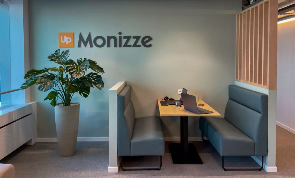 Moderne bedrijfsruimte met Up Monizze-logo, grijze zithoeken, houten tafel met laptop, groene plant en natuurlijke verlichting.