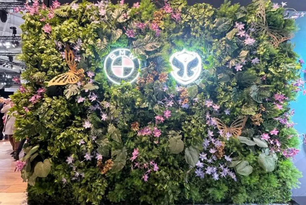 Groene groendecoratie muurtje met roze en paarse bloemen, gouden vlinders en neonverlichting als evenementsdecoratie.