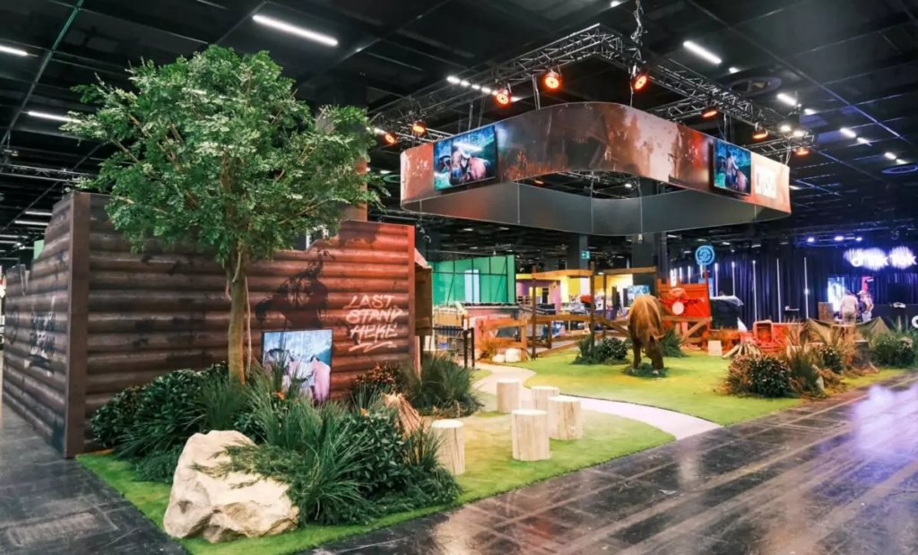 Grote binnenevent met themadecoratie in natuurstijl: houten wanden, kunstbomen, groen gazon, houten logs en verlichte displays in hal.