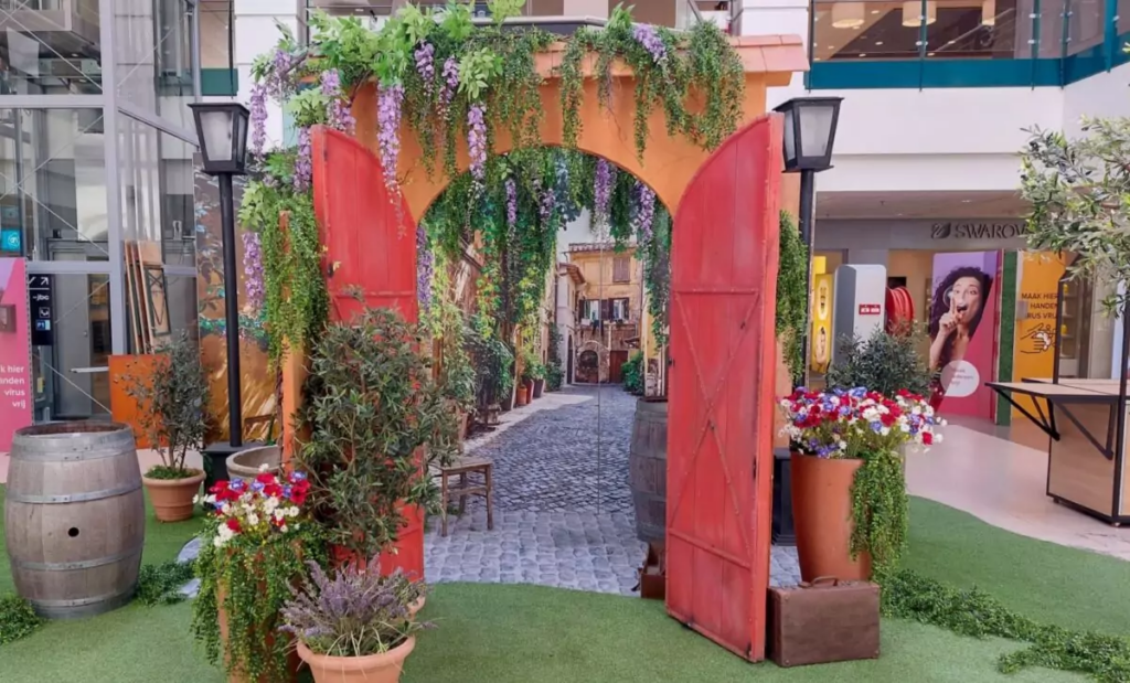 Mediterrane themapoort met rode deuren, hangende paarse bloemen, groene planten en bloempotten in een winkelcentrum.