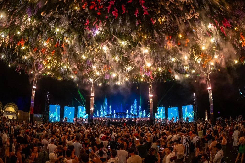 Groot openluchtfestival 's nachts met duizenden bezoekers onder verlichte hangende kunstgroen-installaties in rood, paars en goud.