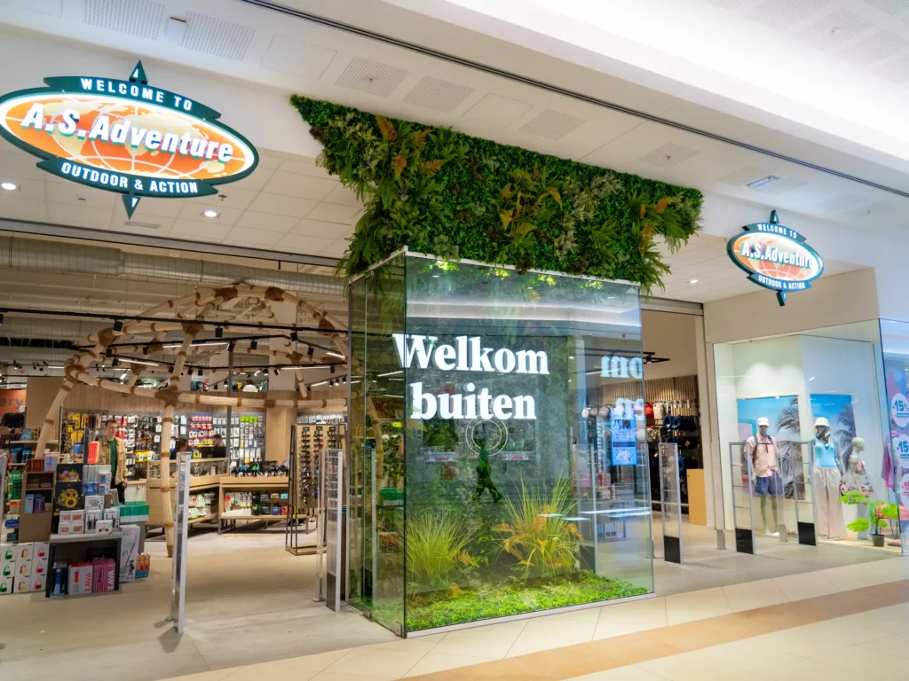A&S Adventure outdoor & action store entrance in a shopping mall with a "Welkom buiten" welcome message on a glass door decorated with living plants.