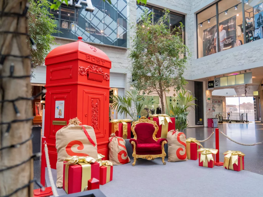 Kerstthema-installatie in een winkelcentrum met rode brievenbus, fluweelstoel, geschenkdozen en groene planten.