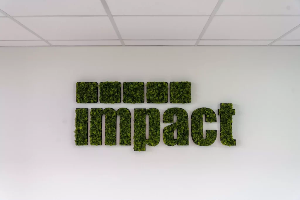 Wandinstallatie met het woord "impact" gemaakt van mos en vijf groene vierkante tegels op een witte muur.