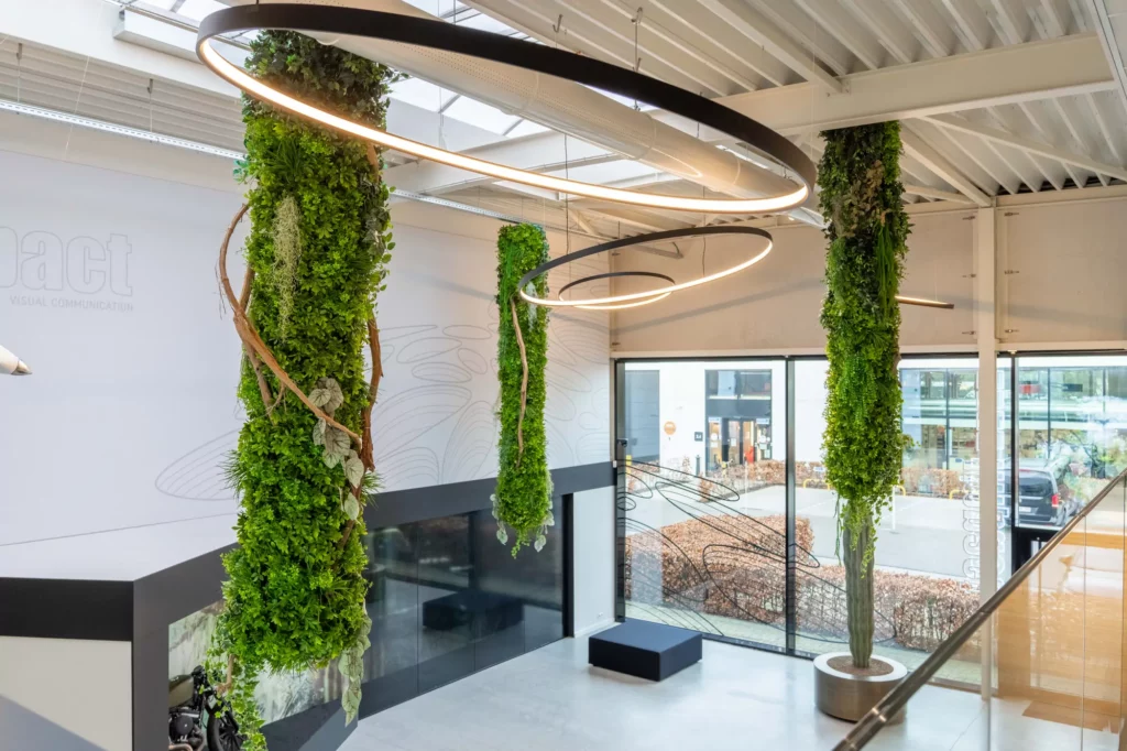 Modern kantoorinterieur met hangende groene planten aan kolommen, organische LED-verlichtingsarmaturen en grote ramen met uitzicht op straat.